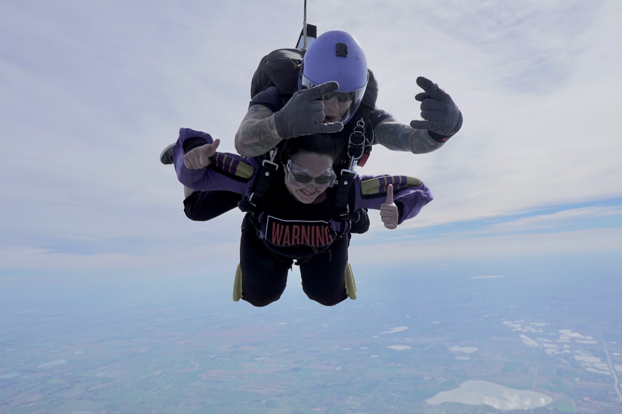 A grinning young woman strapped to her jump‑master gives a double thumbs‑up