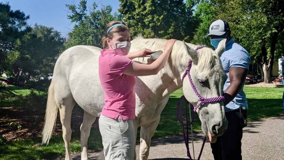 Kelly in sleep shades and mask turns her head to her right while she brushes a white horse named Booger with her left hand. Booger is 