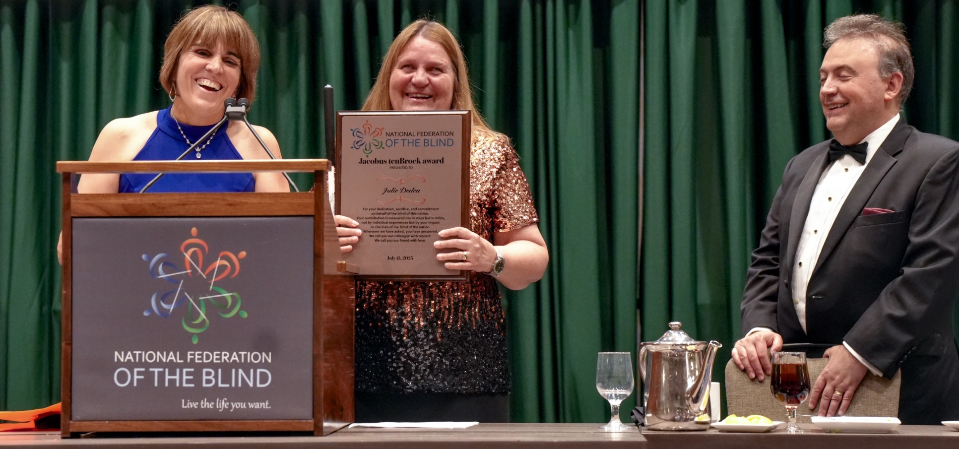 Three smiling people in formal attire; one speaks at a podium, another holds up a plaque, and the third looks on.