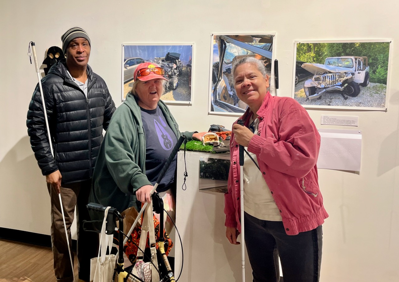 Three blind people with white canes pose before a dramatic piece made by a blind ACC student Shon, Sandy and Saray pose in front of a piece with ceramic and photographs that recreates an automobile accident that caused the artist’s blindness.