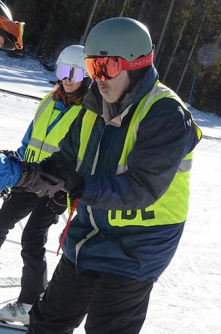 A man wearing a blind skier vest on a snowy slope balances himself on skis with the outstretched hands of a guide while another guide looks on smiling.