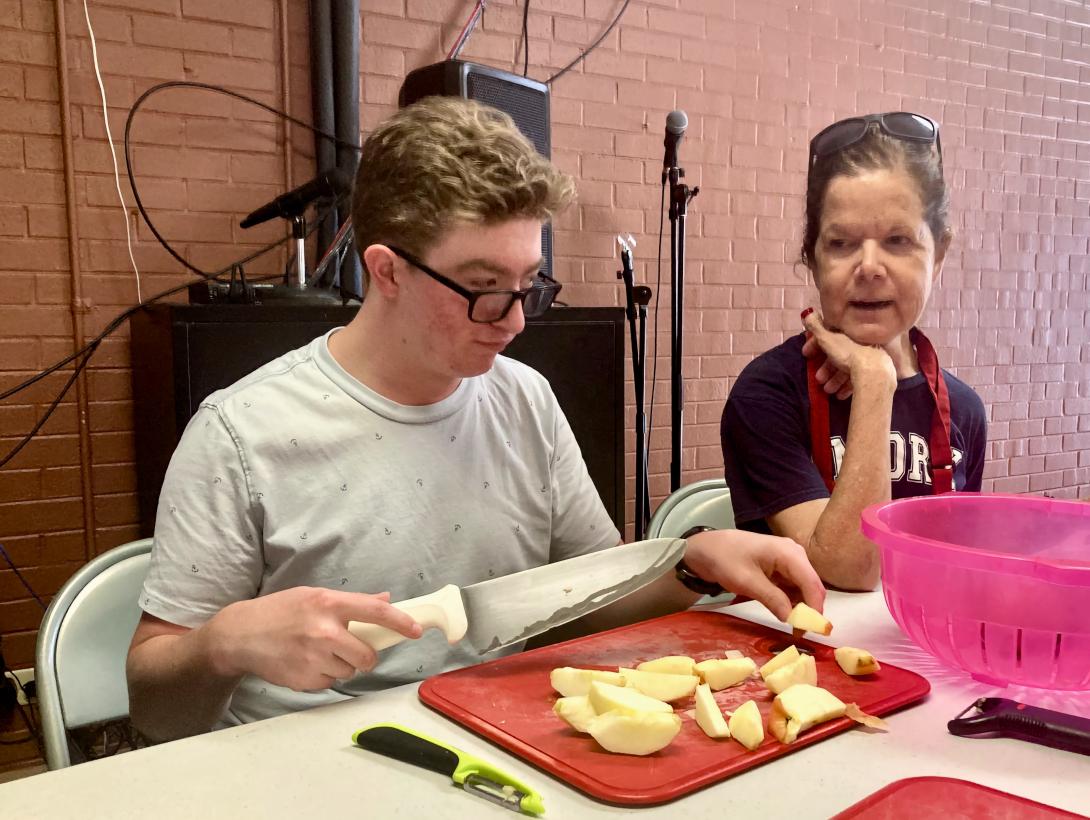 High school student Seth slices apples with a large knife while mentor Heather offers guidance - tangle of cables andconduit on the wall behind Seth's head